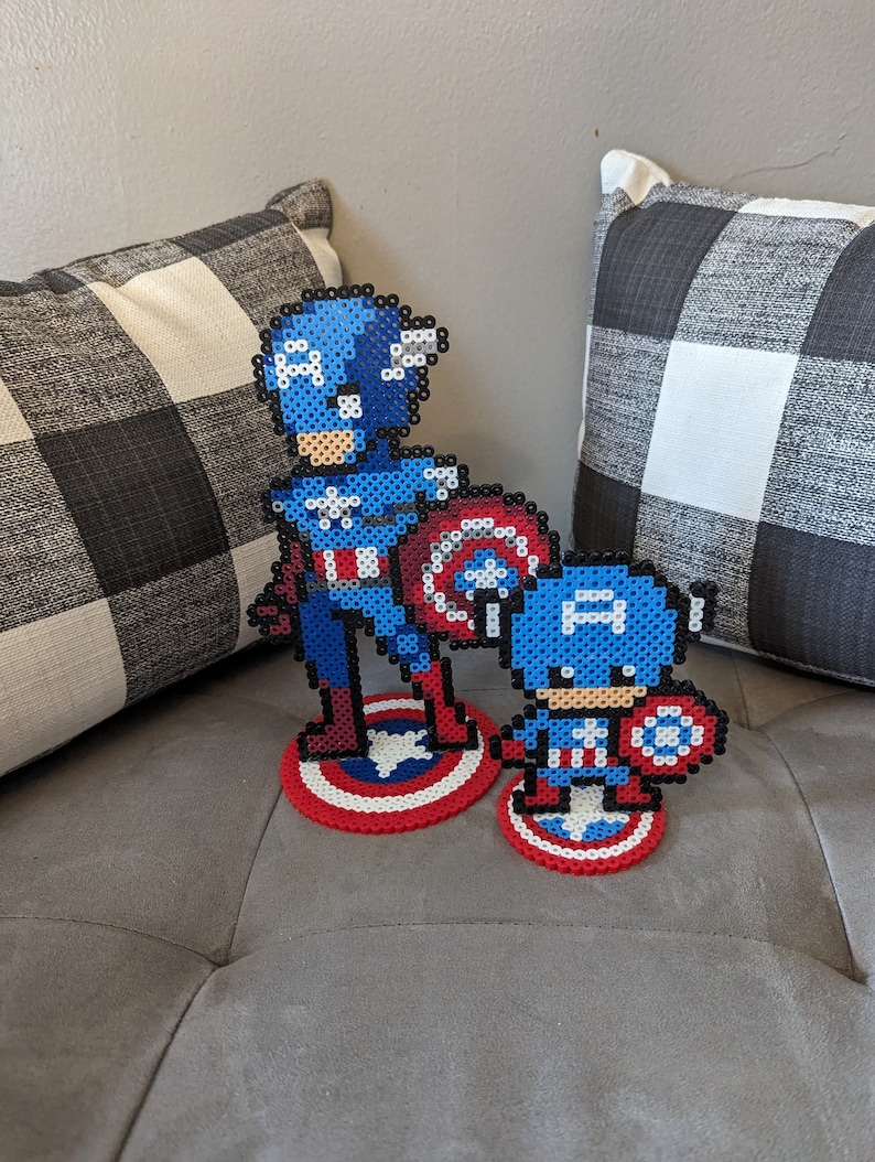 May include: Two Captain America figures made of blue, red, and white perler beads. The larger figure is standing on a red, white, and blue circular base. The smaller figure is standing on a smaller red, white, and blue circular base.