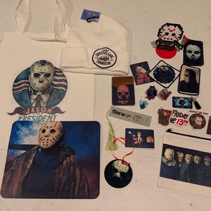 May include: A collection of Friday the 13th themed merchandise including a white tote bag with a Jason President graphic, a white Camp Crystal Lake Counselor beanie, a mousepad, a zipper pouch, various stickers, patches, and a hanging ornament.