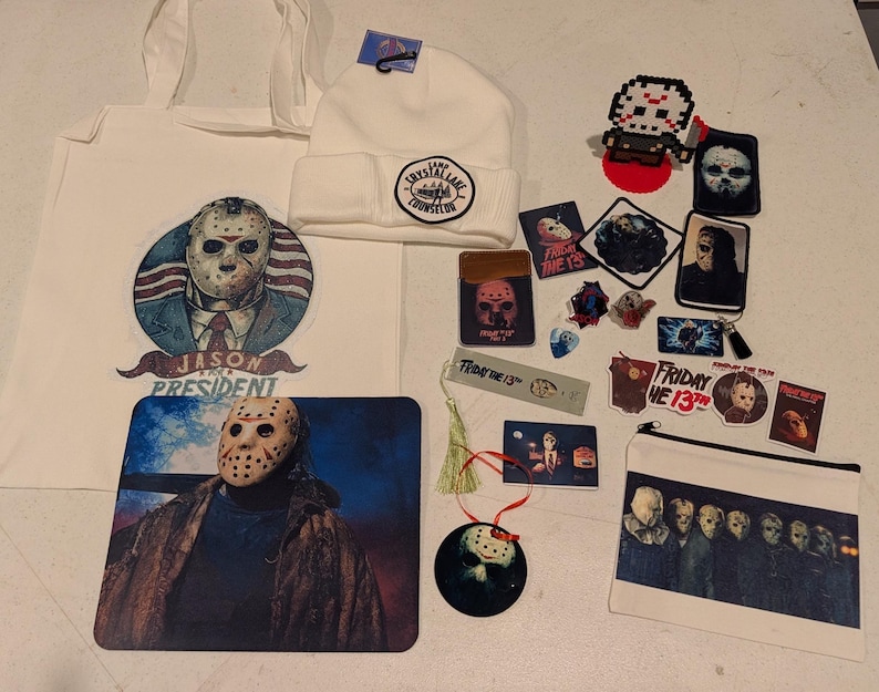 May include: A collection of Friday the 13th themed merchandise including a white tote bag with a Jason President graphic, a white Camp Crystal Lake Counselor beanie, a mousepad, a zipper pouch, various stickers, patches, and a hanging ornament.