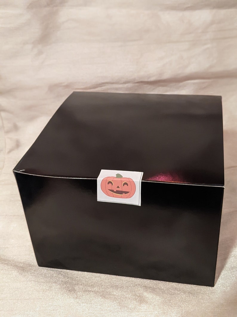 May include: A black gift box with a pumpkin sticker on the top. The pumpkin is orange with a friendly face and a green stem.