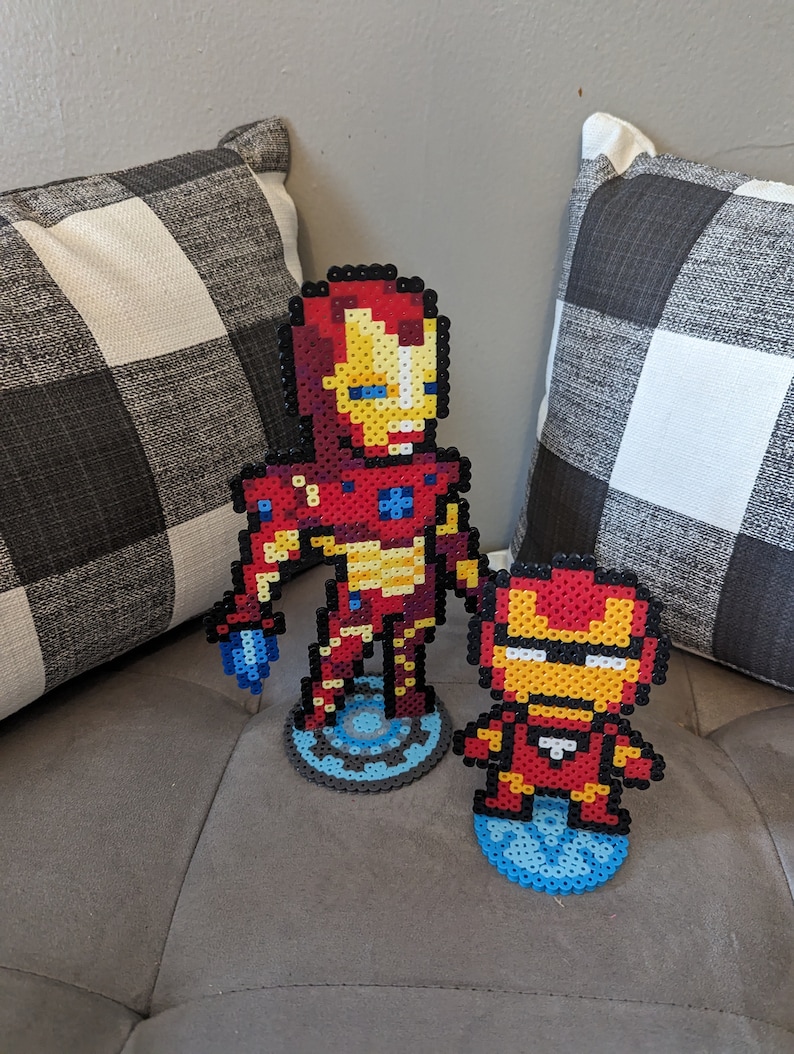 May include: Two Iron Man figures made of red, yellow, and blue perler beads. The larger figure is standing on a blue and gray base with a circular design. The smaller figure is standing on a blue base with a circular design.