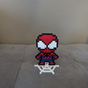 Puede incluir: Un sprite de Spiderman de cuentas rojas y azules de pie sobre una telara&ntilde;a de cuentas blancas.