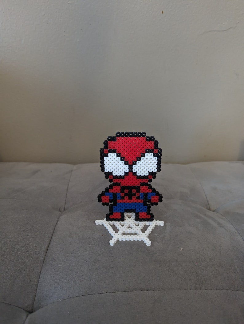 Puede incluir: Un sprite de Spiderman de cuentas rojas y azules de pie sobre una telara&ntilde;a de cuentas blancas.