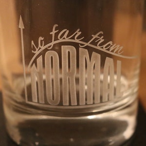 Può includere: Un bicchiere di vetro trasparente con il testo "so far from NORMAL" inciso sul lato. Il testo è in un carattere stilizzato ed è curvo per assomigliare a un grafico.