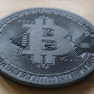 May include: A close-up of a gray, circular, textured Bitcoin coin with the Bitcoin symbol embossed in the center. The coin has the words "BTC" and "Satoshi Nakamoto" inscribed around the edge.