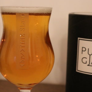 K&ouml;nnte beinhalten: Ein Glas Bier in einem klaren Glas mit einem Etikett, auf dem "Dry Hop" in einer stilisierten Schrift steht. Das Glas steht auf einer Holzoberfl&auml;che neben einer schwarzen Kerze mit dem Text "PURE GLASS" auf dem Etikett.