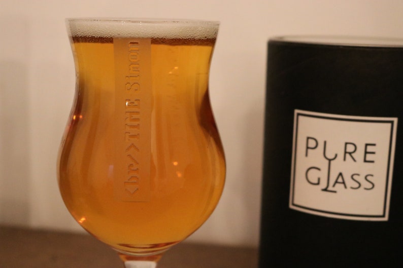 K&ouml;nnte beinhalten: Ein Glas Bier in einem klaren Glas mit einem Etikett, auf dem "Dry Hop" in einer stilisierten Schrift steht. Das Glas steht auf einer Holzoberfl&auml;che neben einer schwarzen Kerze mit dem Text "PURE GLASS" auf dem Etikett.