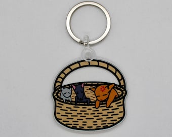 May include: A brown wicker basket keychain with three cartoon cats inside. The cats are grey, black, and orange. The keychain has a silver metal ring.