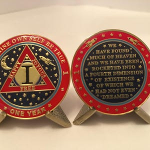 May include: Two commemorative coins with red, blue, and gold accents. One coin features text "TO THINE OWN SELF BE TRUE" and "ONE YEAR". The other coin has the text "WE HAVE FOUND MUCH OF HEAVEN". Both coins are on small metal stands.