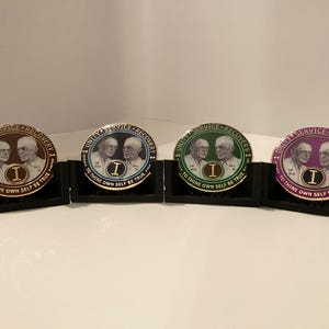 May include: Four gold-colored coins with a white background and black text. The coins feature two men's faces and the text "Unity Service Recovery 1" and "To Thine Own Self Be True". The coins are displayed on black stands.