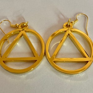 May include: A pair of gold-colored earrings. Each earring features a circle with a triangle inside. The earrings have a hook closure. The jewelry is displayed on a white surface.