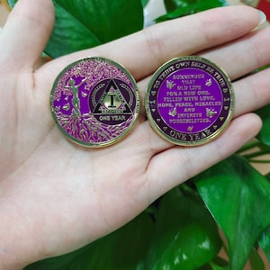May include: Gold coin with a purple center and a gold rim. The front of the coin features a tree with roots and a woman standing in the branches. The back of the coin has text that reads "To thine own self be true. Surrender that old life for a new one, filled with love, hope, peace, miracles and infinite possibilities. One year."