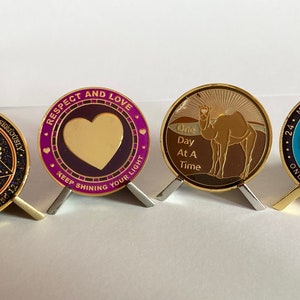 May include: Four gold-colored metal medallions with different designs and text. The first medallion has a dark blue background with a gold triangle and the text "Don't take yourself too damn seriously 62 Rule Page 119 - 122". The second medallion has a purple background with a gold heart and the text "Respect and Love Keep Shining Your Light". The third medallion has a brown background with a gold camel and the text "One Day At A Time". The fourth medallion has a blue background with a gold triangle and the text "24 Miracles Happen Here 24 Unity Service Recovery One Day At A Time".