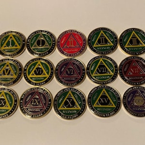 May include: A collection of recovery medallions with gold rims and various colored centers. Each medallion features a triangle with a Roman numeral, the word "RECOVERY", and the number of years. The text "UNITY, SERVICE, RECOVERY" is also present.
