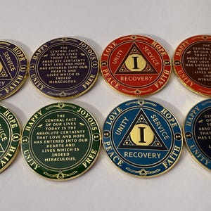 May include: A collection of eight recovery medallions in various colors, including purple, green, red, and blue. Each coin features the letter "I" inside a triangle, surrounded by words like "Love," "Unity," and "Recovery."