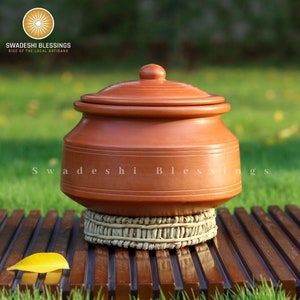 May include: A terracotta cooking pot with a lid, resting on a woven straw mat. The pot is brown and has a smooth, rounded shape. The mat is light brown and has a natural texture. The pot is labelled "Swadeshi Blessings Rise of the Local Artisans".