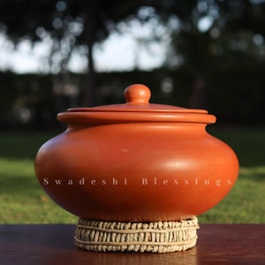 May include: A terracotta pot with a lid, sitting on a woven mat. The pot is a traditional Indian cooking vessel, known as a "handi". The text "Swadeshi Blessings" is printed on the side of the pot.