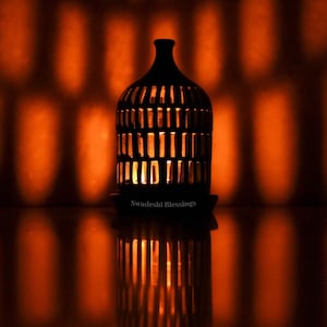 May include: A black metal candle holder with a geometric design, lit with a warm orange glow, reflecting on a dark surface.