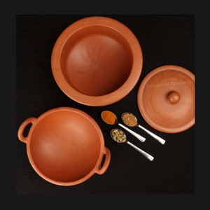 May include: Two terracotta cooking pots with lids, one with a handle, on a black surface.  The pots are surrounded by four spoons with spices, including turmeric, mustard seeds, and cardamom.