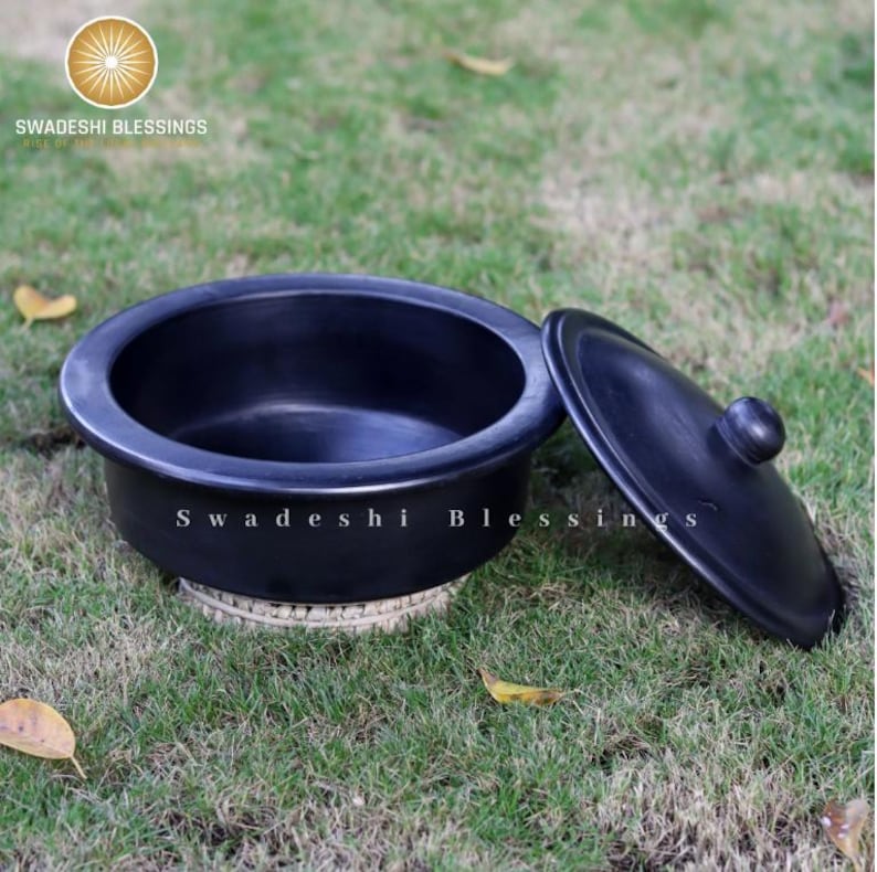 Puede incluir: Una olla de barro negra con tapa, sentada sobre una estera tejida. La olla est&aacute; hecha de barro tradicional indio y se utiliza para cocinar.
