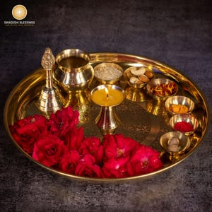 May include: A golden brass puja thali with various items, including a lit candle, small bowls of spices and nuts, a bell, and red roses. The thali is round and has an engraved design. The logo "SWADESHI BLESSINGS" is in the top left corner.