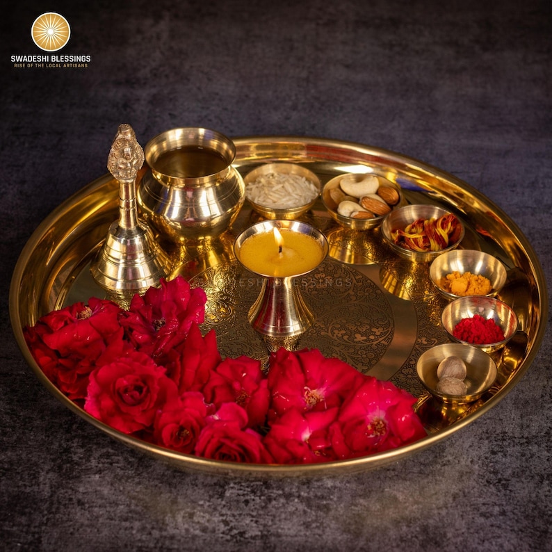 May include: A golden brass puja thali with various items, including a lit candle, small bowls of spices and nuts, a bell, and red roses. The thali is round and has an engraved design. The logo "SWADESHI BLESSINGS" is in the top left corner.