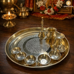 May include: A golden brass puja thali with a lit candle, small bowls of spices and nuts, a bell, and red roses. The thali is round and has an engraved design.