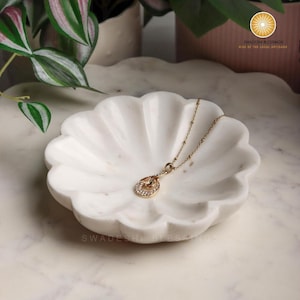 May include: A white marble decorative dish with a scalloped edge, holding a gold necklace with a pendant. The pendant is an oval shape with small, clear stones. The dish is on a marble surface.