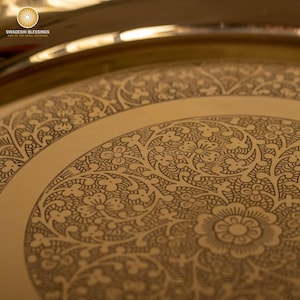 May include: Close-up of a gold-colored decorative tray with intricate floral and leaf patterns etched into the surface. The tray has a wide, circular design with a raised edge. The text "SWADESHI BLESSINGS" is visible in the upper left corner.