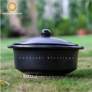 Puede incluir: Olla de barro negro con tapa, sentada sobre una estera tejida. La olla est&aacute; etiquetada con "Swadeshi Blessings".
