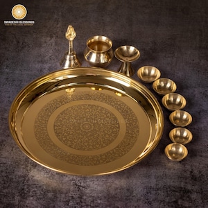 May include: A collection of brass religious items, including a large, round tray with an etched floral design, small bowls, a bell, and other ceremonial pieces. The items are arranged on a dark surface.