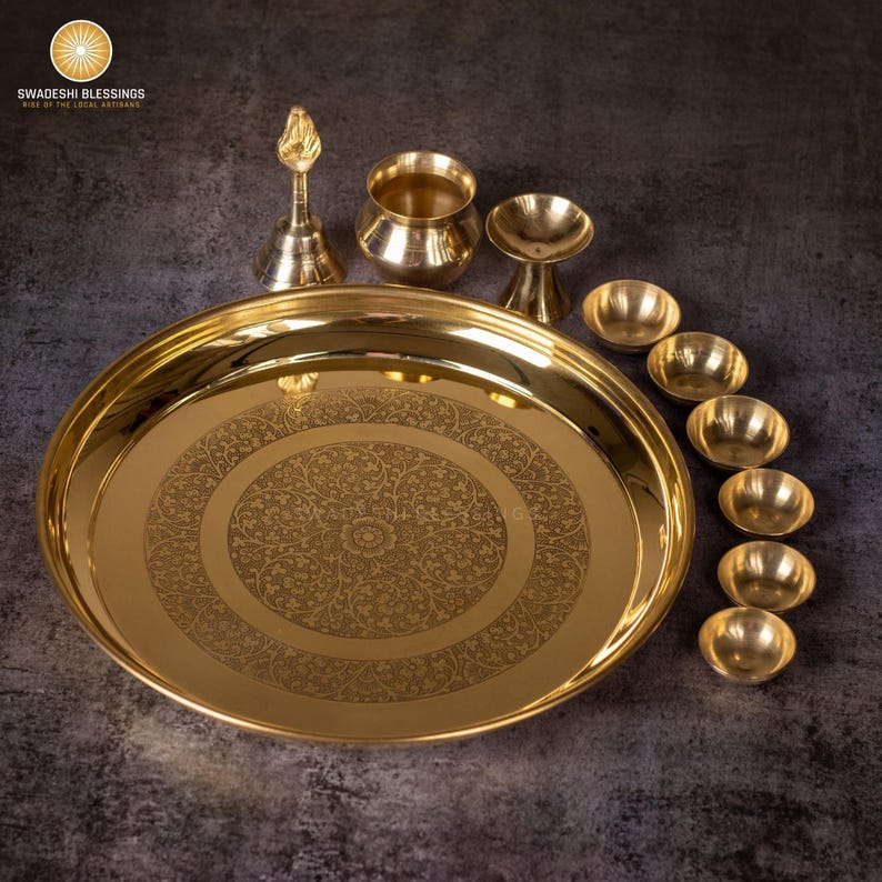 May include: A collection of brass religious items, including a large, round tray with an etched floral design, small bowls, a bell, and other ceremonial pieces. The items are arranged on a dark surface.
