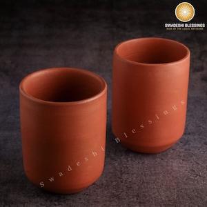 Może przedstawiać: Dwa kubki do picia z terakoty, lub szklanki, z tekstem "Swadeshi Blessings" na boku jednego kubka. Kubki są wykonane z gliny i mają rustykalny, ziemisty wygląd.