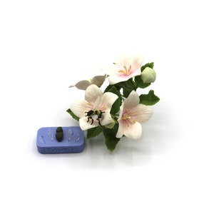 May include: A close-up of a white flower with pink accents, a bee is perched on the flower. The flower is part of a small bouquet of white flowers with green leaves. A blue silicone mold with five impressions is in the background.