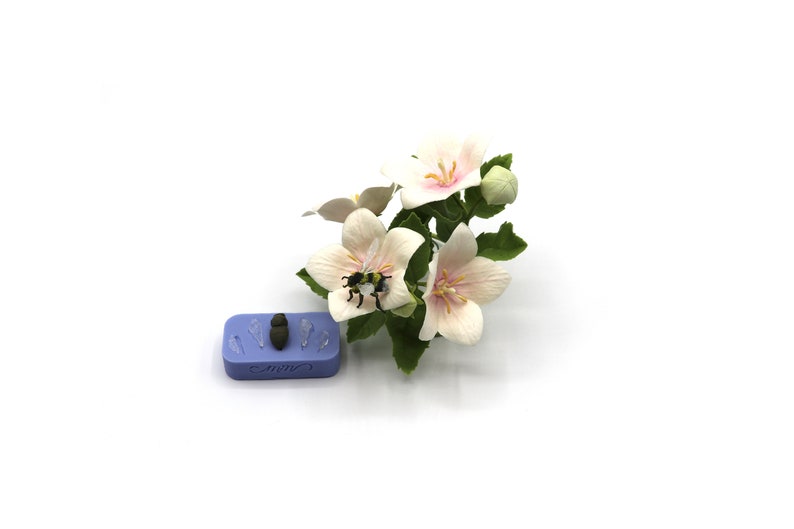 Pu&ograve; includere: Primo piano di un fiore bianco con accenti rosa, un'ape &egrave; posata sul fiore. Il fiore fa parte di un piccolo bouquet di fiori bianchi con foglie verdi. Uno stampo in silicone blu con cinque impressioni &egrave; sullo sfondo.