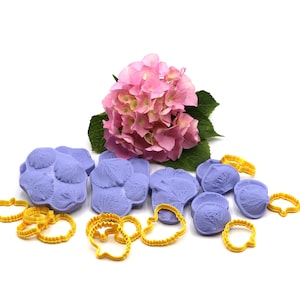 Könnte beinhalten: Ein Satz violetter Silikonformen in Form von Hortensienblütenblättern, mit einer rosa Hortensie im Hintergrund. Die Formen sind von gelben Kunststoff-Ausstechern umgeben.