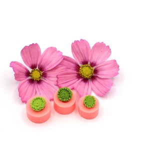 May include: Three pink and green silicone moulds for making resin crafts. The moulds are shaped like flowers with a round centre and petals. The moulds are sitting on a white surface.