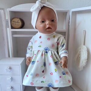May include: A doll wearing a white dress with a floral pattern, a matching headband, and white booties. The dress features a variety of colorful flowers and small insects. The doll is positioned in front of a white cabinet.