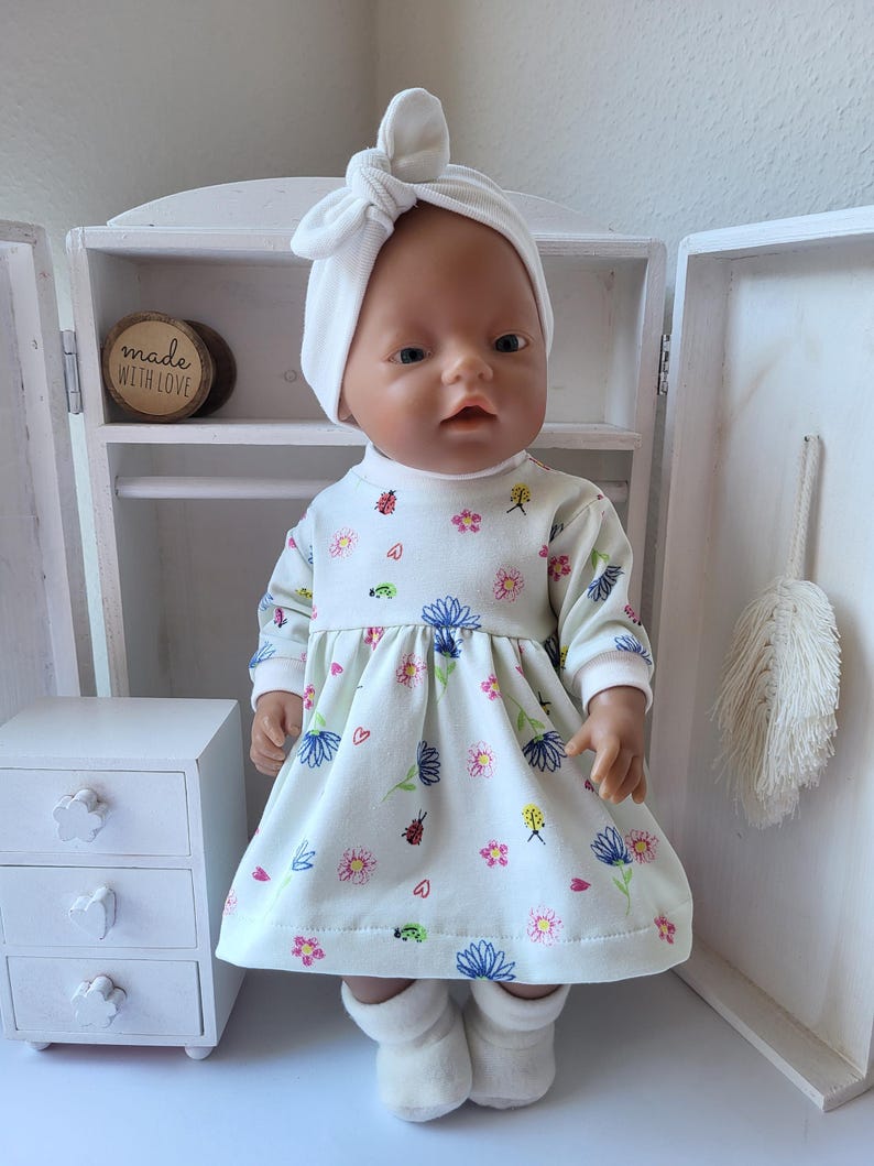 May include: A doll wearing a white dress with a floral pattern, a matching headband, and white booties. The dress features a variety of colorful flowers and small insects. The doll is positioned in front of a white cabinet.