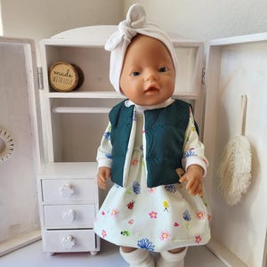 May include: A doll wearing a white head wrap, a dark green vest, and a floral print dress. The doll is standing in front of a white cabinet with a small wooden sign that says "made with love". The dress has a pattern of flowers and ladybugs.