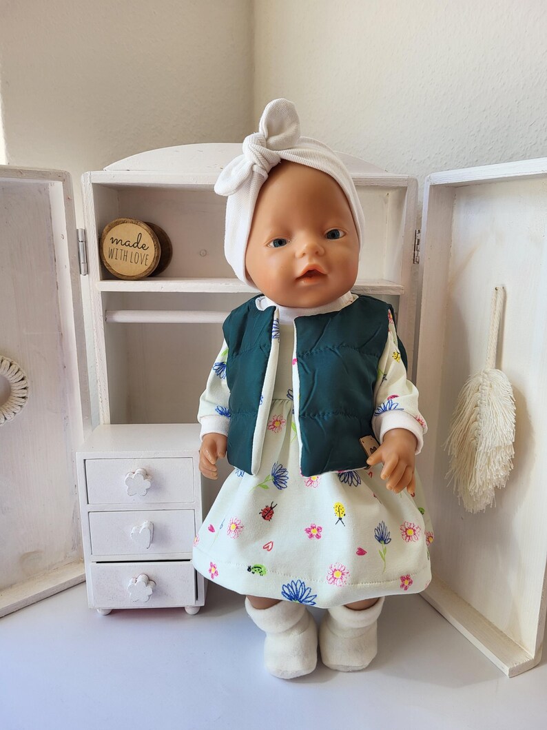 May include: A doll wearing a white head wrap, a dark green vest, and a floral print dress. The doll is standing in front of a white cabinet with a small wooden sign that says "made with love". The dress has a pattern of flowers and ladybugs.