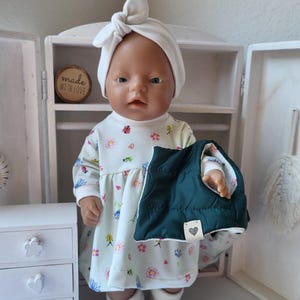 May include: A doll wearing a white dress with a floral pattern, a white head wrap, and white socks. The doll is holding a dark green quilted vest. A small wooden sign reads "made with love."