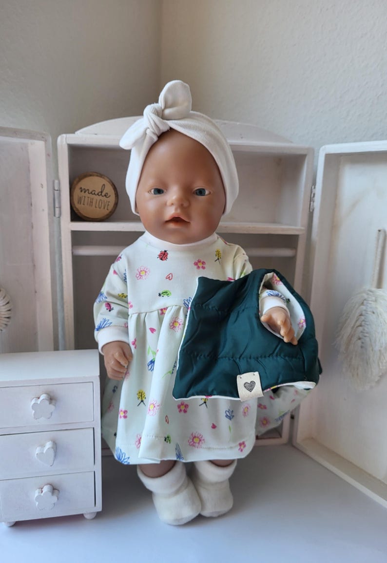 May include: A doll wearing a white dress with a floral pattern, a white head wrap, and white socks. The doll is holding a dark green quilted vest. A small wooden sign reads "made with love."