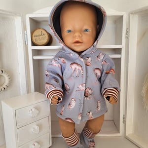 Könnte beinhalten: Eine Puppe in einem hellblauen Kapuzen-Outfit mit einem Waldtiermuster. Das Outfit umfasst eine Jacke, Socken und Babyschuhe. Die Jacke hat braun-weiß gestreifte Bündchen. Links befindet sich ein kleiner weißer Schrank mit blumen- und herzförmigen Schubladengriffen.