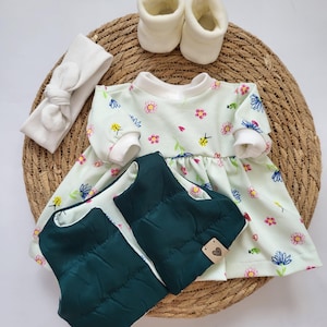 May include: A baby outfit set featuring a light green floral print dress, a dark green quilted vest, a white headband, and white booties. The items are arranged on a woven mat, creating a charming ensemble for a newborn.