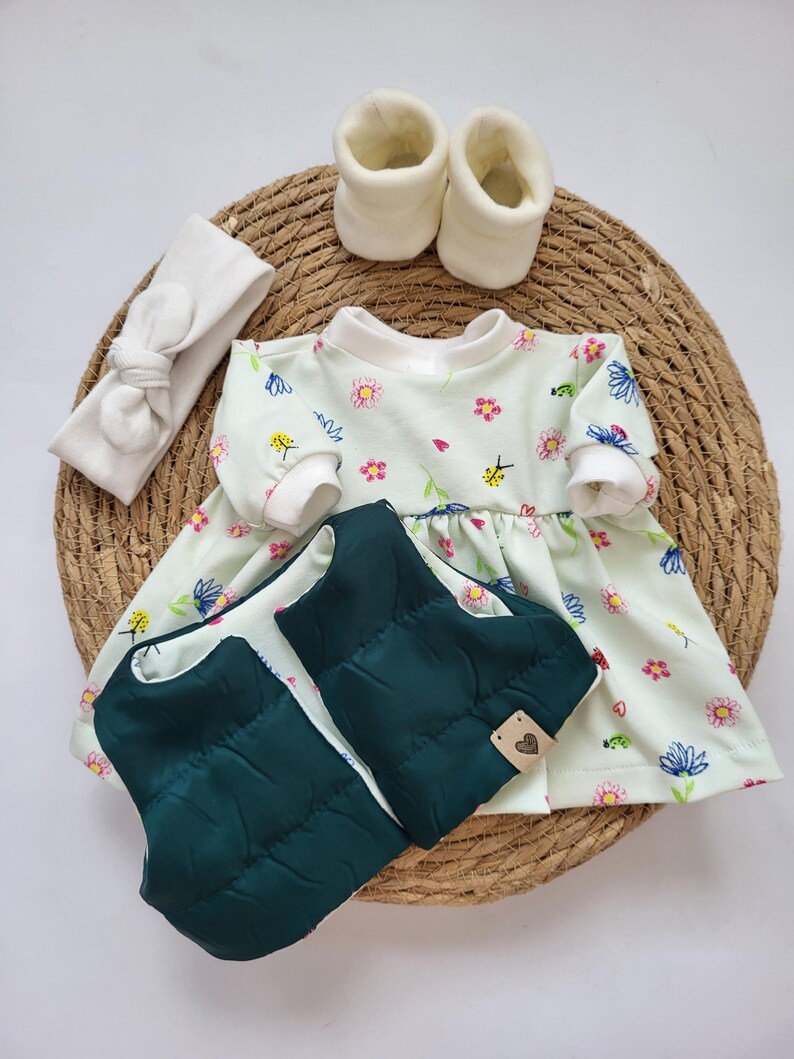 May include: A baby outfit set featuring a light green floral print dress, a dark green quilted vest, a white headband, and white booties. The items are arranged on a woven mat, creating a charming ensemble for a newborn.