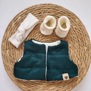 May include: A dark green baby vest with a white floral lining, a white headband with a bow, and two white baby booties. The vest has a small tag with a heart. The items are arranged on a woven, circular mat.
