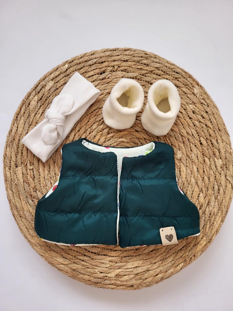 May include: A dark green baby vest with a white floral lining, a white headband with a bow, and two white baby booties. The vest has a small tag with a heart. The items are arranged on a woven, circular mat.