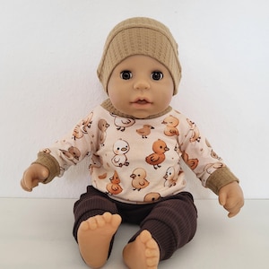 Könnte beinhalten: Eine Puppe mit einer beigefarbenen Strickmütze, einem langärmeligen Shirt mit Entenmuster und einer dunkelbraunen Ripphose. Das Shirt hat einen hellrosa Hintergrund mit Cartoon-Enten. Die Puppe sitzt mit ausgestreckten Beinen.