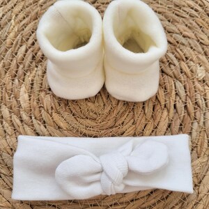 May include: A set of baby accessories, including two ivory-colored booties and a matching headband with a bow. The items are made of a soft, knitted material and are displayed on a woven, light brown surface.